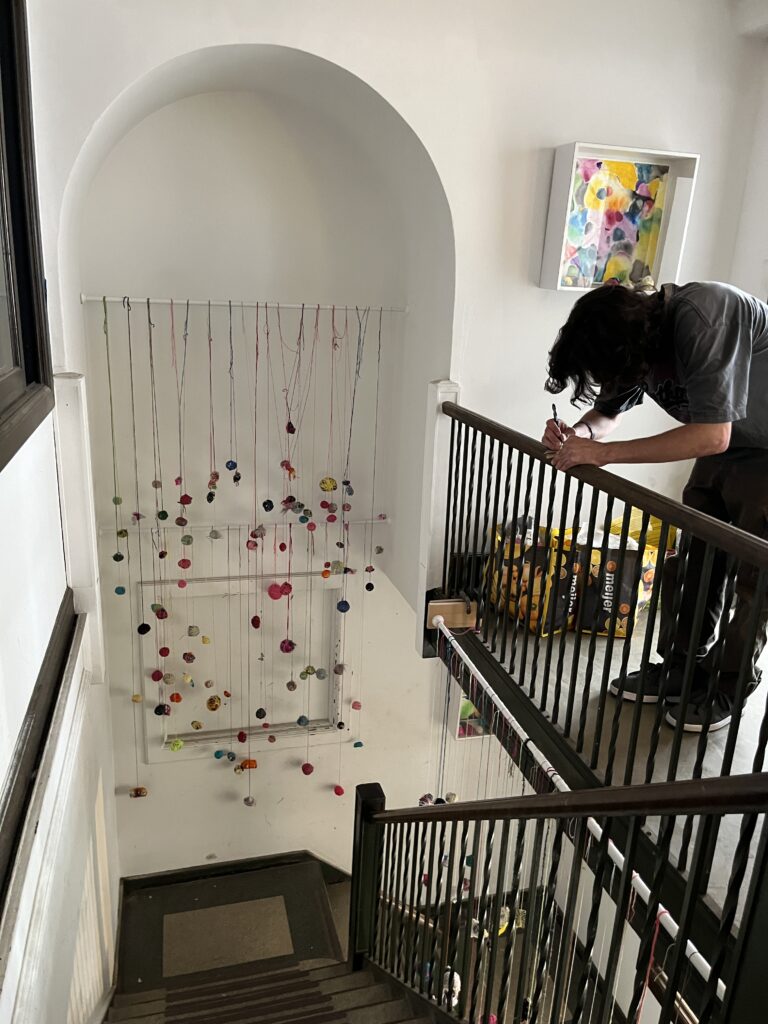 Bundles of Joy installation by Kelly Darke at the Environment + Microclimates show 2024 by Fiber Club Detroit at Boyer Campbell Building, Detroit, MI during Detroits Month of Design. Image shows installation hanging in stairwell and alcove as well as individual participating by making a bundle of joy meditation.